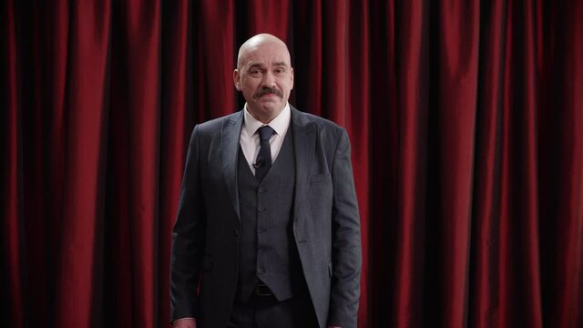 Late-night talk show host is performing his monologue against red curtain, looking into camera, looking into camera. TV broadcast style show. Shot with RED cinema camera