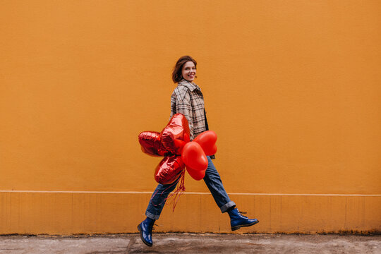 Full-length Portrait Of Positive Short-haired Girl In Jeans, Boots Dr. Martins And Tweed Jacket. Woman With Red Balloons Walks Against Tbackground Of Orange Wall