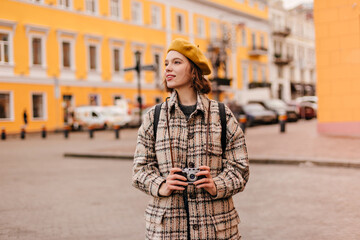 Obraz premium Pretty girl in bright beret looks at urban architecture with enthusiasm. Lady in stylish autumn coat holding retro camera