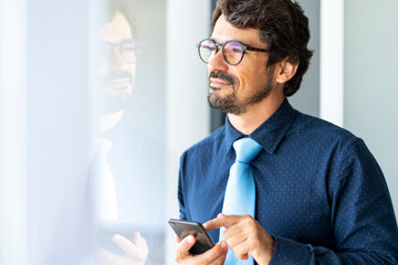 Businessman wearing glasses looking through window with the phone on his hands. Successful male...