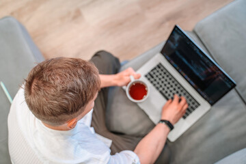 Obraz premium Top view of a young male programmer drinking coffee and writing on a laptop keyboard. The concept of people working with technology and computer at home