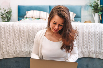 A young woman uses a laptop while lying on the bed. The concept of the home office and online education.