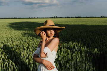 Beautiful young woman in a boho style dress and hat on a field with wheat at sunset.