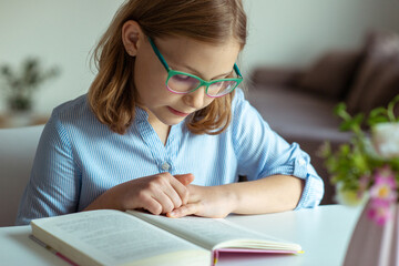 Pretty teen girl reading book at desk during pandemic distance learning