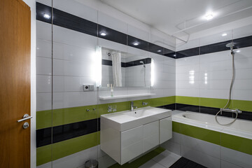 Contemporary interior of bathroom in modern flat. Black and green tile. Sink and mirror. Bath with shower.