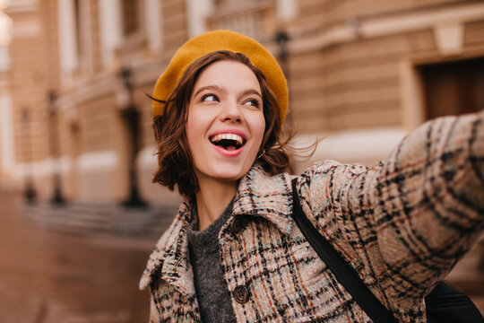 Close-up portrait of emotional Parisian girl in coat and beret. Woman with smile makes selfie on background of cityscape