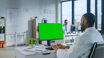 Black Medical Doctor is Working on a Computer with Green Screen Mock Up Display in a Health Clinic. Assistant in White Lab Coat is Reading Medical History Behind a Desk in Hospital Office.