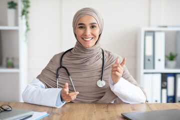 Smiling female doctor sitting at workdesk in clinic, making consultation