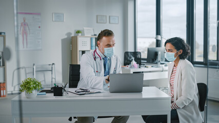 Family Doctor in Protective Mask is Reading Medical History of Female Patient and Speaking with Her During Consultation in a Health Clinic. Physician in Lab in Front of Computer in Hospital Office. 