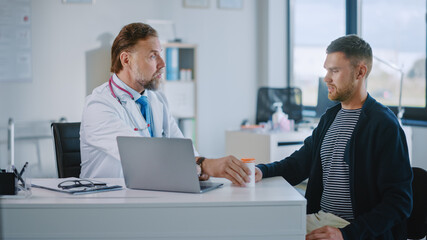 Fototapeta premium Senior Doctor is Prescribes Medicine in a Box to a Male Patient. Medical Consultation in a Health Clinic. Physician in White Lab Coat Sitting Behind a Computer in Hospital Office. 