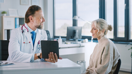 Fototapeta premium Friendly and Cheerful Family Doctor is Reading Medical History of Senior Female Patient During Consultation in a Health Clinic. Physician Using Tablet Computer in Hospital Office.