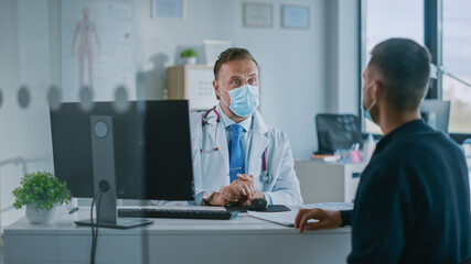 Family Doctor in Protective Mask is Reading Medical History of Young Male Patient and Speaking with Him During Consultation in a Health Clinic. Physician in Lab in Front of Computer in Hospital Office