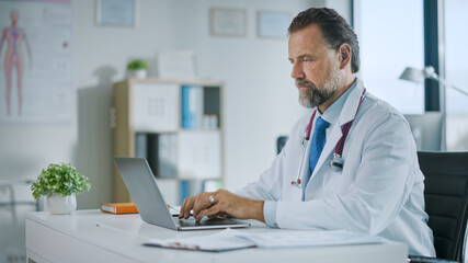 Happy Family Medical Doctor is Working on a Laptop Computer in a Health Clinic. Physician in White Lab Coat is Browsing Medical History Behind a Desk in Hospital Office. 