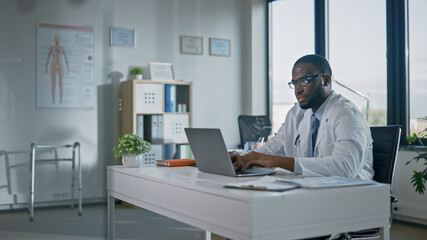 Calm African American Family Medical Doctor in Glasses is Working on a Laptop Computer in a Health Clinic. Physician in White Lab Coat is Browsing Medical History Behind a Desk in Hospital Office. 