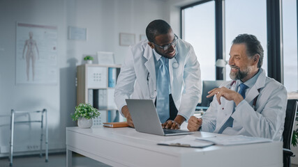 Fototapeta premium Young African American Physician is Talking to a Senior Doctor in a Medical Health Clinic. Colleagues Discuss Work on Laptop Computer in Hospital Office. Assistant Asks Medical Advice from a Doctor.