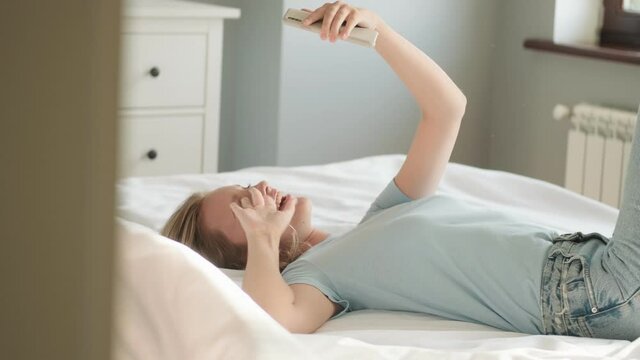 A smiling woman is talking with a video connection using her smartphone lying in her bed in the morning