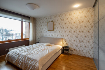Contemporary interior of bedroom in modern flat. Cozy bed. Lamps on nightstands. Huge window. Hardwood floor.