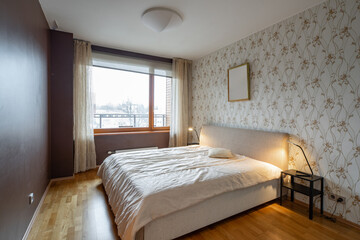 Contemporary interior of bedroom in modern flat. Cozy bed. Lamps on nightstands. Huge window. Hardwood floor.