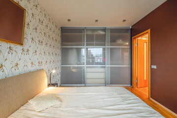 Contemporary interior of bedroom in luxury flat. Side view of cozy bed. Huge sliding door wardrobe. Lamp on nightstand.