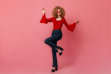Naughty girl in pink sunglasses is having fun and dancing on isolated background. Photo of blonde wearing red blouse, dark pants and high heels