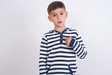 little cute boy kid wearing stripped t-shirt against white wall being in stupor shocked, has astonished expression pointing at oneself with finger saying: Who me?