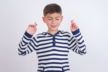 Joyful little cute boy kid wearing stripped t-shirt against white wall clenches teeth, raises fingers crossed, makes desirable wish, waits for good news, I have to win.
