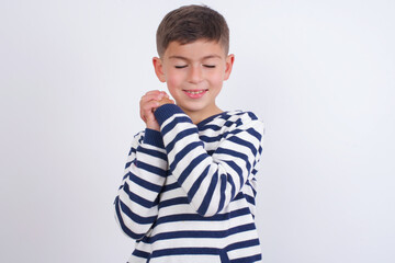 Dreamy little cute boy kid wearing stripped t-shirt against white wall with pleasant expression, closes eyes, keeps hands crossed near face, thinks about something pleasant