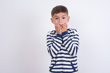 Anxiety - little cute boy kid wearing stripped t-shirt against white wall covering his mouth with hands scared from something or someone bitting nails.