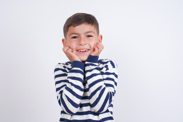 Dreamy little cute boy kid wearing red stripped t-shirt against white wall keeps hands pressed together under chin, looks with happy expression, has toothy smile.