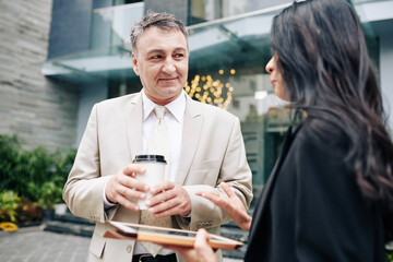 Smiling senior entrepreneur listening to creative ideas of his assistant when they are drinking coffee outdoors