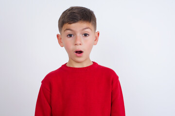 Emotional attractive Little cute boy kid wearing red knitted sweater against white with opened mouth expresses great surprise and fright, stares at camera. Unexpected shocking news and human reaction.
