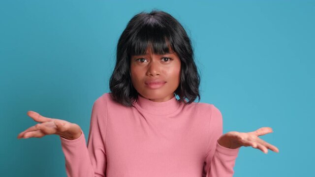 Close up of puzzled dark skinned woman spreads her arms and asks what she can do to solve the problem. Woman in pink clothes posing on a blue background. Concept of negative human emotions.
