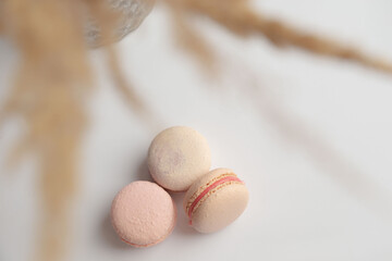 Three tender French macaroon lie on a white background under a bouquet of flowers. Place for an inscription. Dessert. Macaroon. Confectioner. Biscuit. Gift. Postcard. View from above.