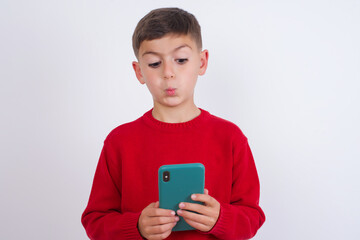 Little cute boy kid wearing red knitted sweater against white wall looks with bugged eyes, holds modern smart phone, receives unexpected message from friend, reads reminder.