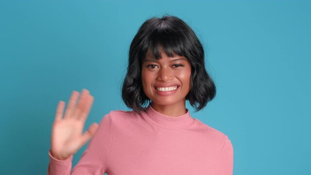 Friendly young Asian woman with dark skin waves her hand and greets smiling broadly at camera meeting new people standing on blue background. Concept of blogging, vlogging and positive emotions.