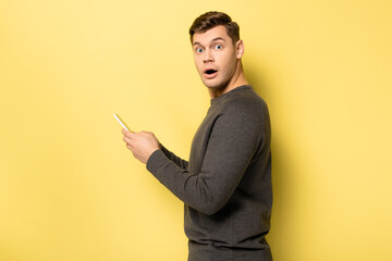 Amazed man looking at camera while using smartphone on yellow background