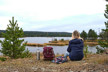 woman is sitting in front of a river, forest and little island. backpack, plaid, thermos are near her. Spruce is on the background. Solo female tourism. Lifestyle moment.