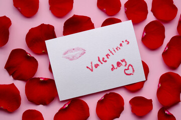 Different red petals of roses and postcard with mark of lips and lettering valentine's day on pink background