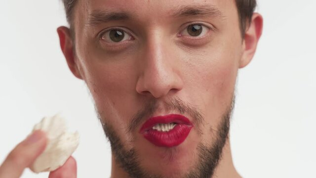 One confident handsome man with beard, moustache, wear makeup – red lipstick on lips, look closely at camera, eat delicious sweet marshmallow, takes a bite, chews isolated on white background close-up