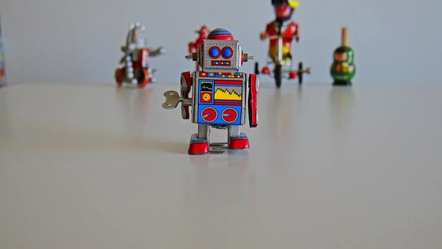 Funny colorful wind-up toy metal robot walks from the center to the camera slowly with others toys behind. Full HD 1080p. Still frame.