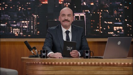 Late-night talk show host sitting behind his table and performing his monologue, looking into camera. TV broadcast style show