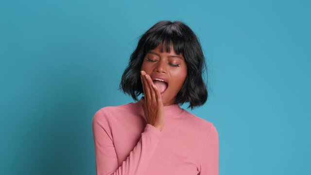 Sleepy young dark skinned woman yawns with her mouth wide open against blue background female closes her mouth with hand thinking about sleep. Concept of jet lag fatigue or well being after party.