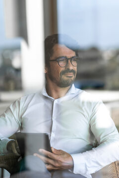 Businessman Wearing Glasses Sitting Down Holding His Tablet And Looking Through The Window. Successful Male Portrait With Positive Attitude Working From Home