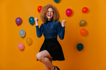 Active girl in black skirt and blue blouse makes winning gesture and laughs, posing on yellow background with balloons