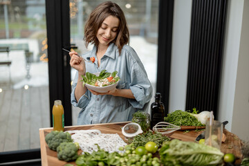 Portrait of young woman smiling and eating healthy green salad with tomato at home. Health care and...