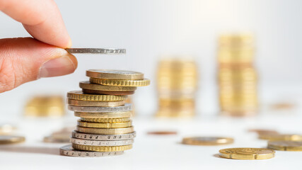 Hand adding coin on pile of euros with others stacked in background. Finger holding metallic cash...