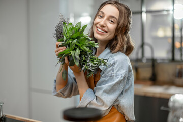 Portrait of cheerful woman in apron with fresh spicy herbs basil, rosemary, thyme on the kitchen....