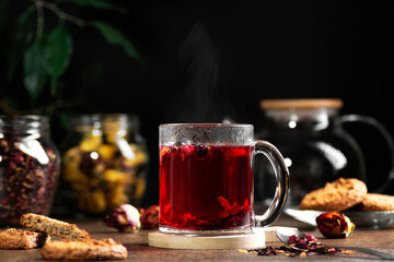 Transparent mug with hot red tea on a dark background, near cookies and tea leaves, horizontal orientation