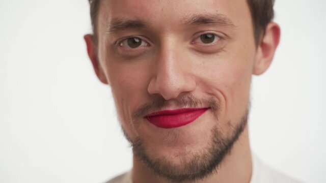 Close up face of happy pleased handsome brunette bearded man use makeup – red lipstick on lips. Caucasian guy stare at camera, smiling isolated on white background. Facial view of young fashion male