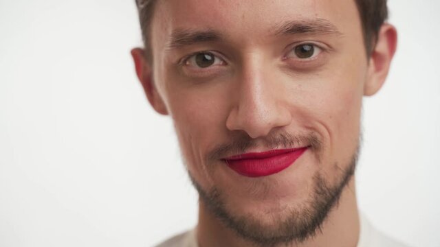 Single young happy pleased man with thick beard, moustache wear make-up – red lipstick on lips, winks isolated on white wall background. Close up face portrait of fashion metrosexual or gay model guy.